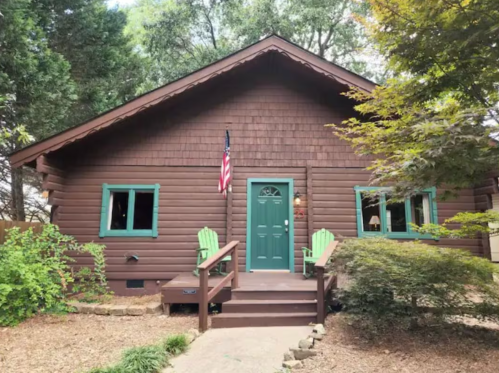 A cozy brown log cabin with green trim, featuring a front porch and an American flag, surrounded by trees.