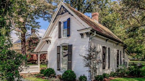 A charming white house with blue shutters, surrounded by greenery and trees, featuring a sloped roof and decorative trim.