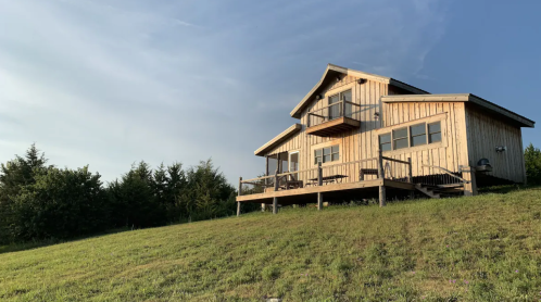A wooden house on a grassy hill, featuring a deck and surrounded by trees under a clear blue sky.