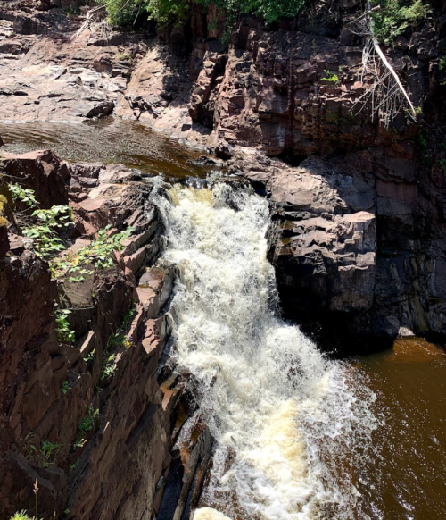 A waterfall cascading over rocky terrain, surrounded by lush greenery and a dark pool below.