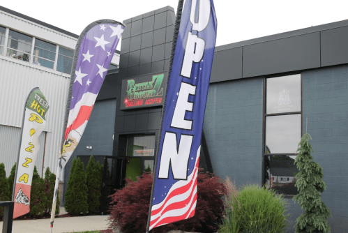 A storefront with "OPEN" flags and patriotic designs, featuring greenery and a pizza sign outside.
