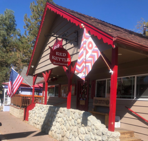 Exterior of the Red Kettle Restaurant featuring red accents, flags, and a stone wall in a wooded area.