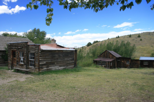 Two rustic wooden buildings sit on a grassy area, surrounded by trees and rolling hills under a blue sky with clouds.