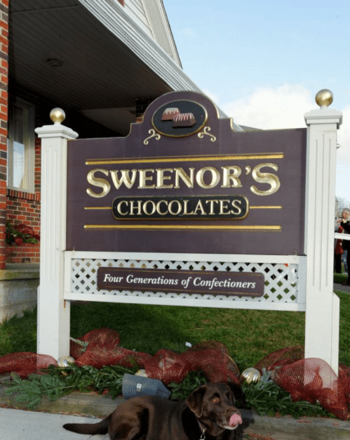 Sign for Sweeney's Chocolates with a dog in front, featuring "Four Generations of Confectioners" below the name.