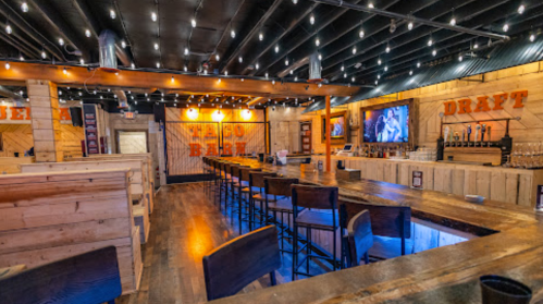 Interior of a rustic bar with wooden decor, a long counter, high stools, and string lights overhead.