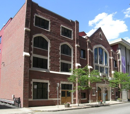 A brick building with large windows and decorative elements, surrounded by trees and a clear blue sky.