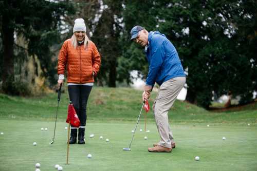 A man putts on a golf green while a woman watches, both dressed for cool weather. Golf flags and balls are scattered around.