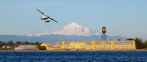 A seaplane flies over a lake with a yellow hotel and a water tower, set against a backdrop of snow-capped mountains.