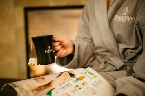 A person in a gray robe holds a black mug while reading a magazine by a cozy fireplace.