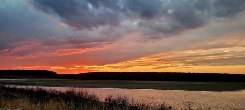 A vibrant sunset over a calm river, with colorful clouds reflecting in the water and a silhouette of trees in the background.