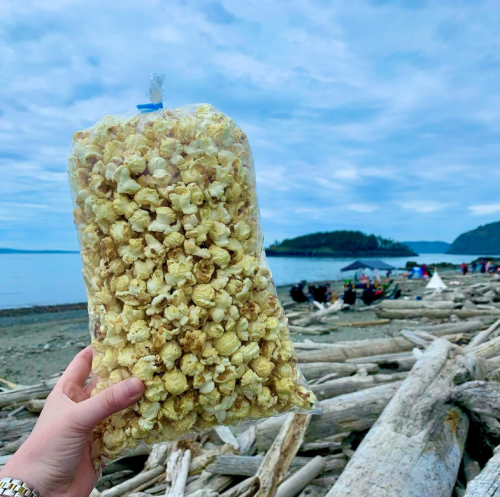A hand holds a large bag of popcorn against a scenic beach backdrop with people and driftwood in the distance.
