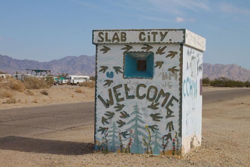 A colorful welcome sign for Slab City, featuring painted birds and flowers, set against a desert landscape.