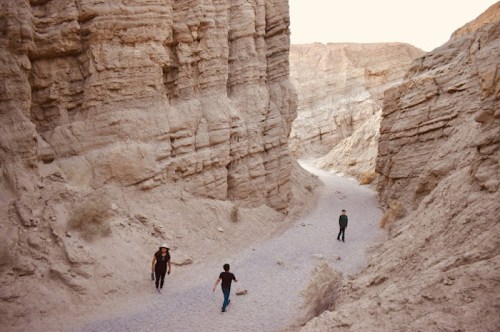 Three people walking through a narrow, rocky canyon with steep, textured walls and a gravel path.