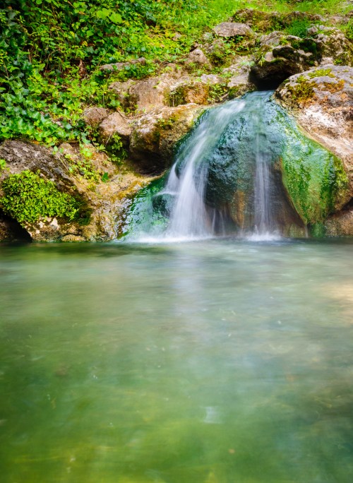 A serene waterfall cascades over mossy rocks into a clear, tranquil pool surrounded by lush greenery.