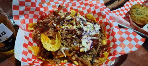 A plate of nachos topped with pulled pork, barbecue sauce, and coleslaw, served with a beer in the background.