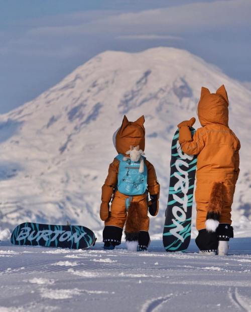 Two children in fox costumes stand on snow, holding snowboards, with a snowy mountain in the background.
