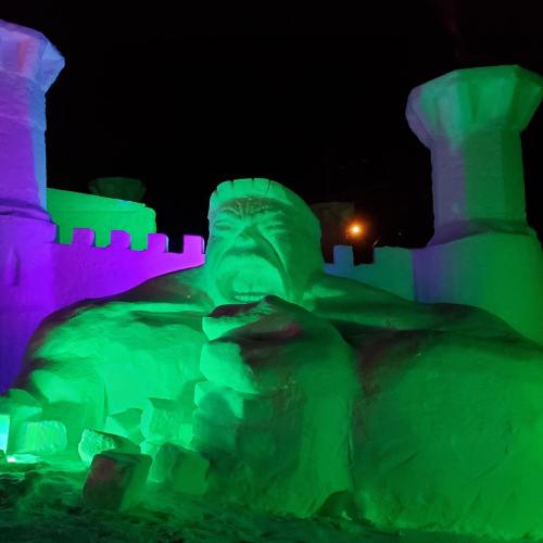 A large, illuminated snow sculpture of a monster, surrounded by colorful lights and a castle-like structure at night.