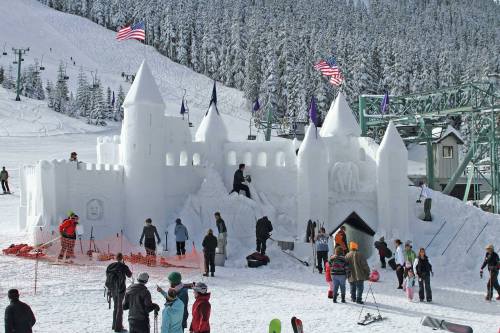 A large snow castle with flags, surrounded by people enjoying winter activities in a snowy landscape.