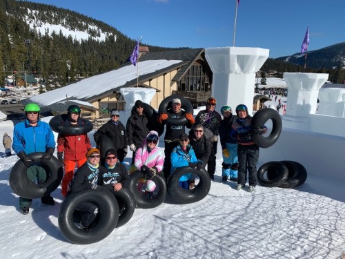 A group of people in winter gear poses with inner tubes on a snowy hill, surrounded by trees and a building in the background.
