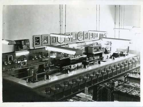 A vintage black-and-white photo of a diner interior, featuring a long counter with stools and a menu on the wall.