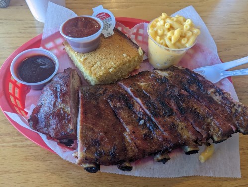A plate of BBQ ribs with macaroni and cheese, cornbread, and two cups of sauce on a red-checkered tray.