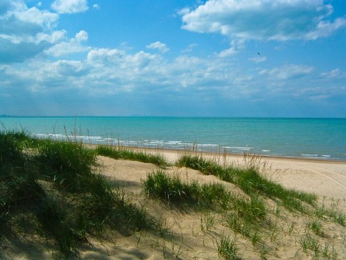 A serene beach scene with soft sand dunes, green grass, and a calm turquoise sea under a partly cloudy sky.