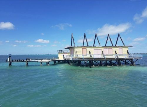 A yellow building on stilts over turquoise water, with a wooden pier extending towards the horizon under a blue sky.