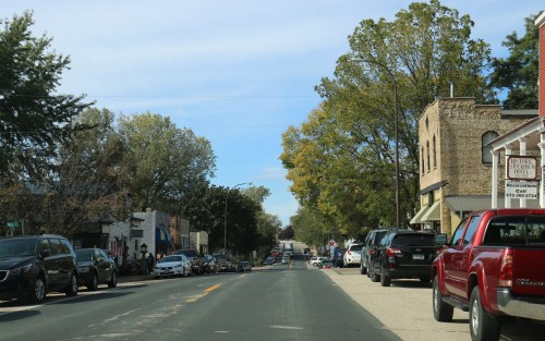 A view of a small town street lined with trees, shops, and parked cars under a clear blue sky.