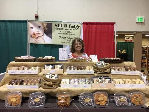 A vendor smiles behind a table filled with various fudge and cookies at a market, with a banner in the background.