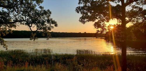 Sunset over a calm lake, framed by trees, with golden light reflecting on the water's surface.
