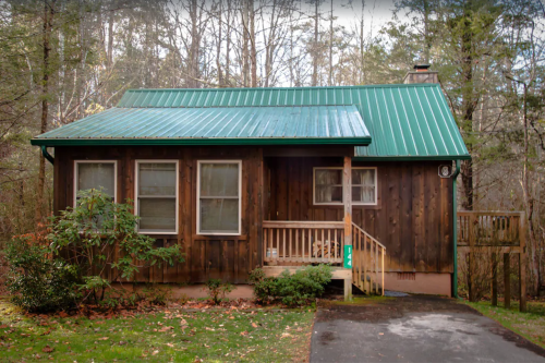 A wooden cabin with a green metal roof, surrounded by trees and a small front porch.
