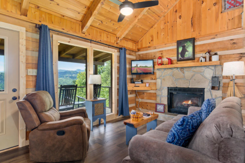 Cozy cabin interior with wooden walls, a stone fireplace, comfortable seating, and scenic mountain views through large windows.
