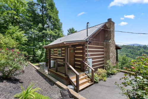 A charming log cabin surrounded by greenery, featuring a stone chimney and a welcoming front porch.