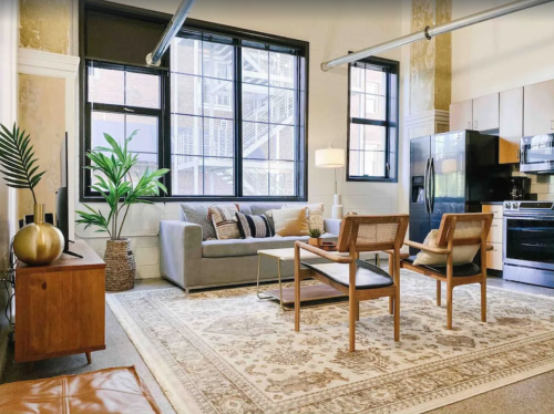 Cozy living room with a gray sofa, two chairs, a coffee table, and large windows, featuring plants and modern decor.