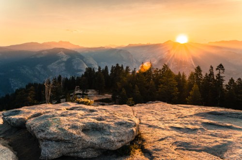 Sunset over a mountainous landscape, with golden rays illuminating the peaks and a forest in the foreground.