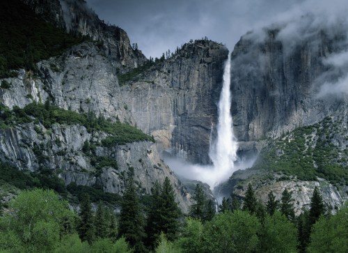 A majestic waterfall cascades down rocky cliffs, surrounded by lush greenery and misty clouds in a mountainous landscape.