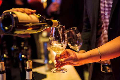 A hand pours white wine into a glass held by another person at a social event, with bottles in the background.