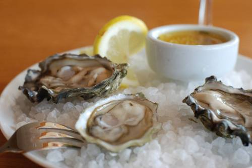 A plate of fresh oysters on ice, garnished with lemon slices and a small bowl of dipping sauce.
