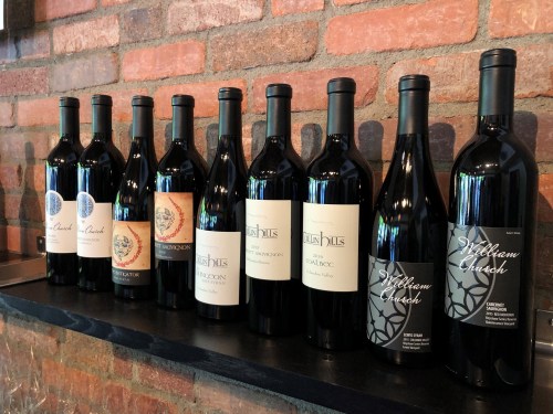 A row of wine bottles on a shelf against a brick wall, featuring various labels and designs.