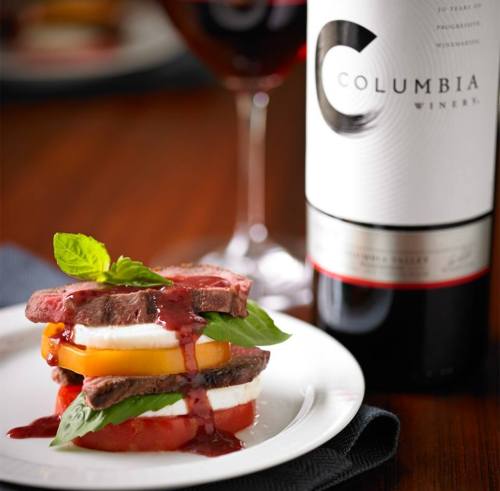 A gourmet dish of layered steak, tomatoes, and basil, served with a glass of Columbia wine in the background.