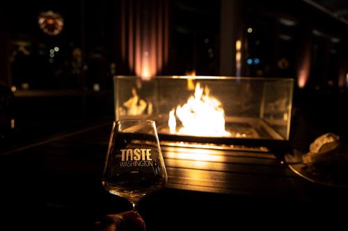 A hand holding a wine glass in front of a glowing fire pit, with "Taste Washington" etched on the glass.