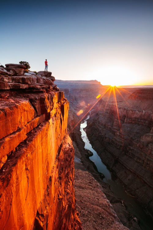 A person stands on a rocky cliff at sunset, overlooking a winding river in a canyon with vibrant orange hues.