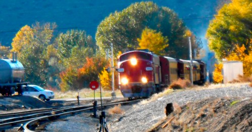 A red train approaches a railway crossing surrounded by colorful autumn trees and a clear blue sky.
