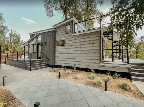Modern wooden house with large windows, a deck, and landscaped pathway surrounded by trees.