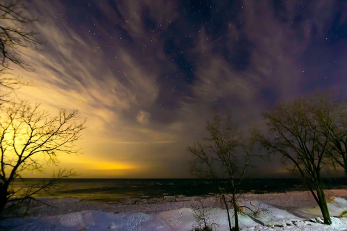 A serene winter night by the lake, with snow-covered ground, silhouetted trees, and a starry sky illuminated by clouds.
