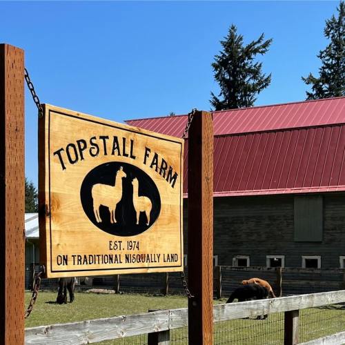 Sign for Topstall Farm, established in 1974, located on traditional Nisqually land, with a red barn in the background.