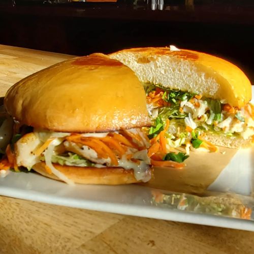 A close-up of a sandwich with a golden bun, filled with shredded vegetables and chicken, served on a white plate.