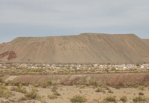 A barren landscape with a large, sloped hill in the background and small houses in the foreground, under a cloudy sky.