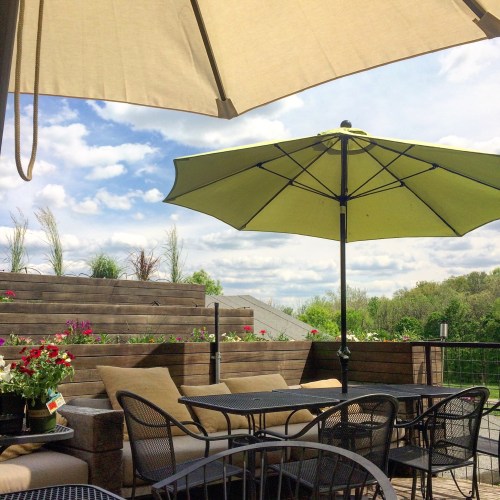A patio with a green umbrella over a table and chairs, surrounded by greenery and a cloudy sky.