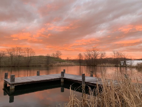 A serene lake at sunset, with a wooden dock and vibrant orange clouds reflecting on the water.
